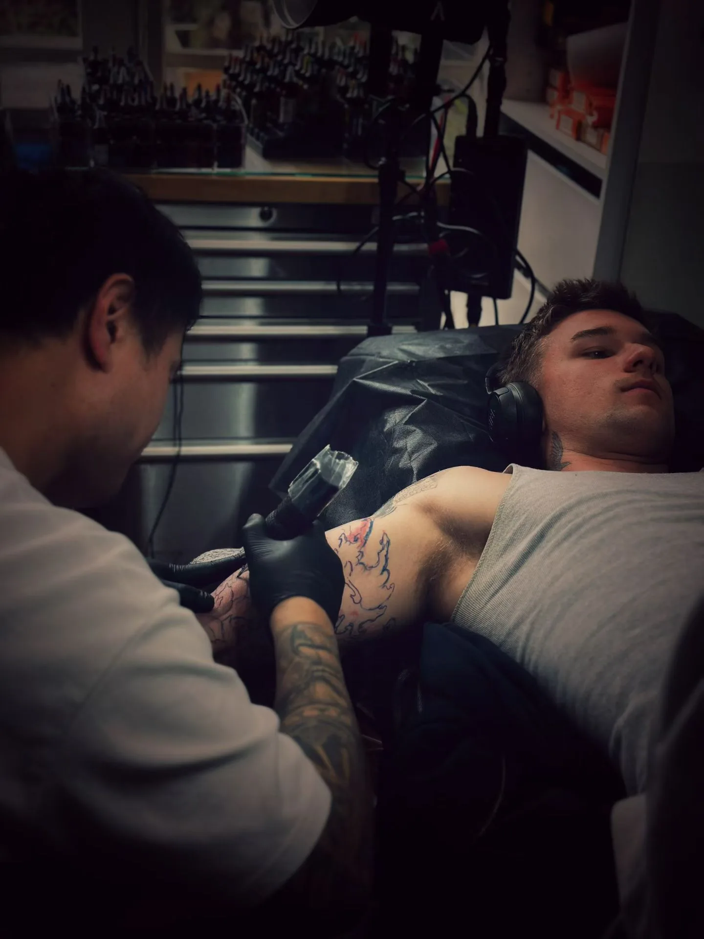 A live tattoo session under warm lamplight — a gloved artist working a reclining client's arm, ink bottles on the wall behind