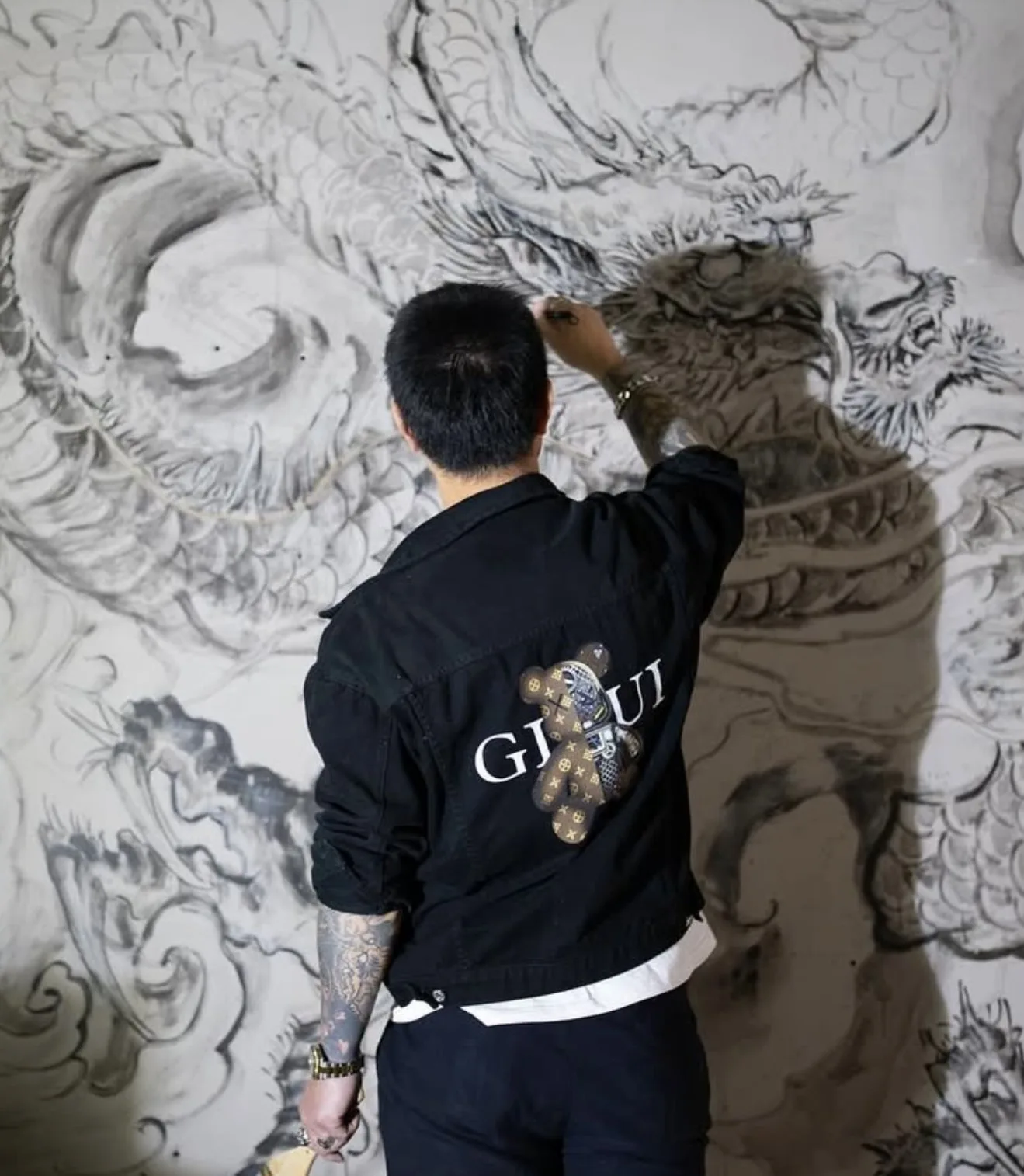 An artist hand-drawing a large Chinese ink-wash dragon mural on the studio wall, back to camera